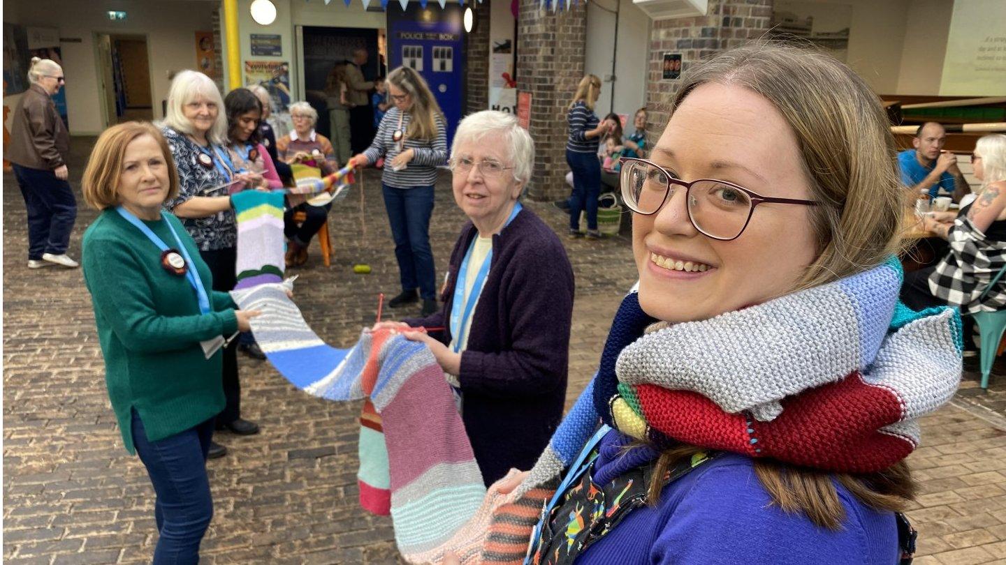 A photo of a group of women holding up the very long scarf and smiling at the camera