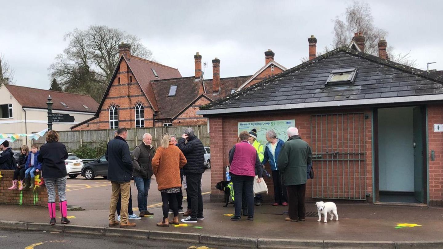 Lewell Street toilets in Newent. It is a a small rectangular red brick building, situated on the corner of a car park next to the road. There is a large group of people standing outside wearing coats and wellies.