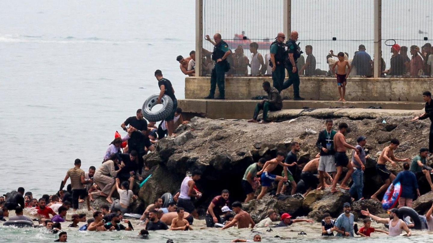 A group of people scramble from the sea up the rocks; several are still in the water with flotation devices. At the top of the rock is a wire fence.