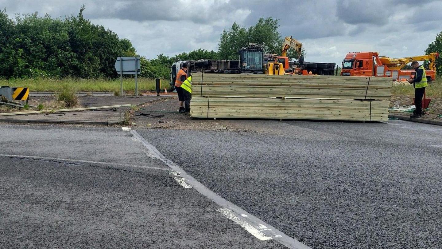 Overturned lorry on the road- with wooden planks in front of the lorry and two men in High-visibility jacket standing on either side of the wooden planks 