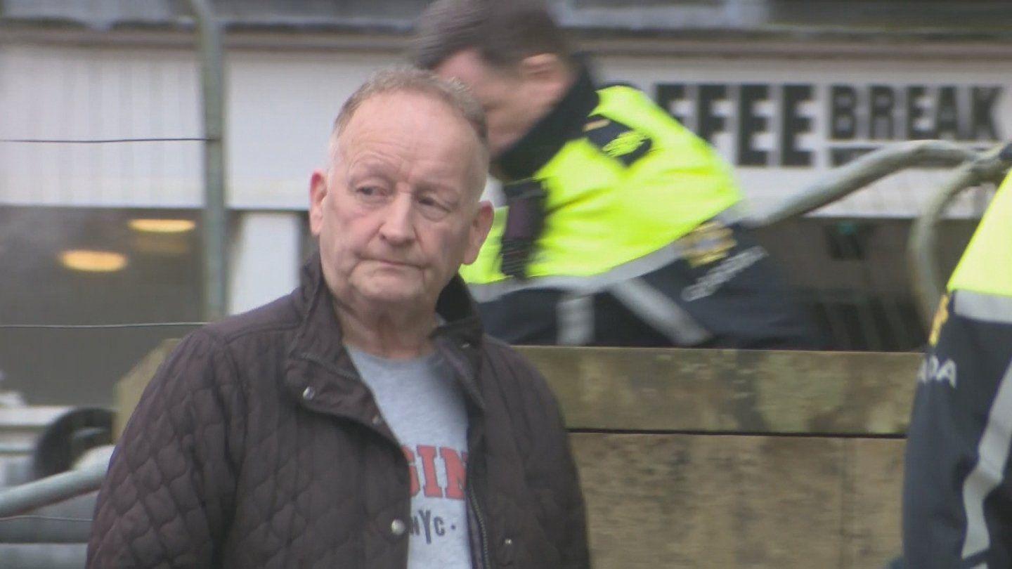 Anthony McGinn stands in front of a wall, with two irish police officers in the background. He is wearing a grey t-shirt and a brown court. 