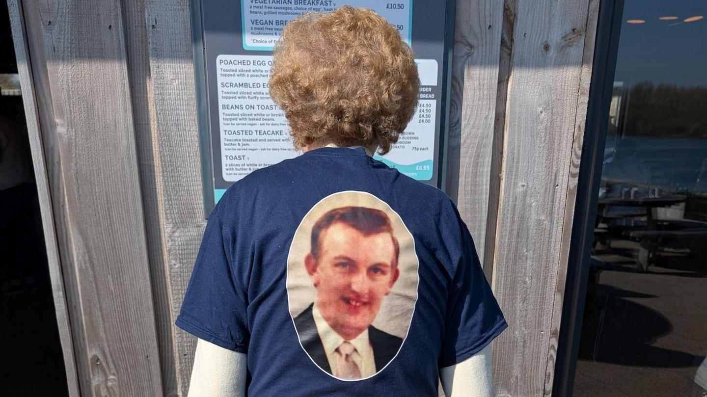 A woman is standing with her back to the camera. She has a blue T-shirt on with a picture of her late husband.