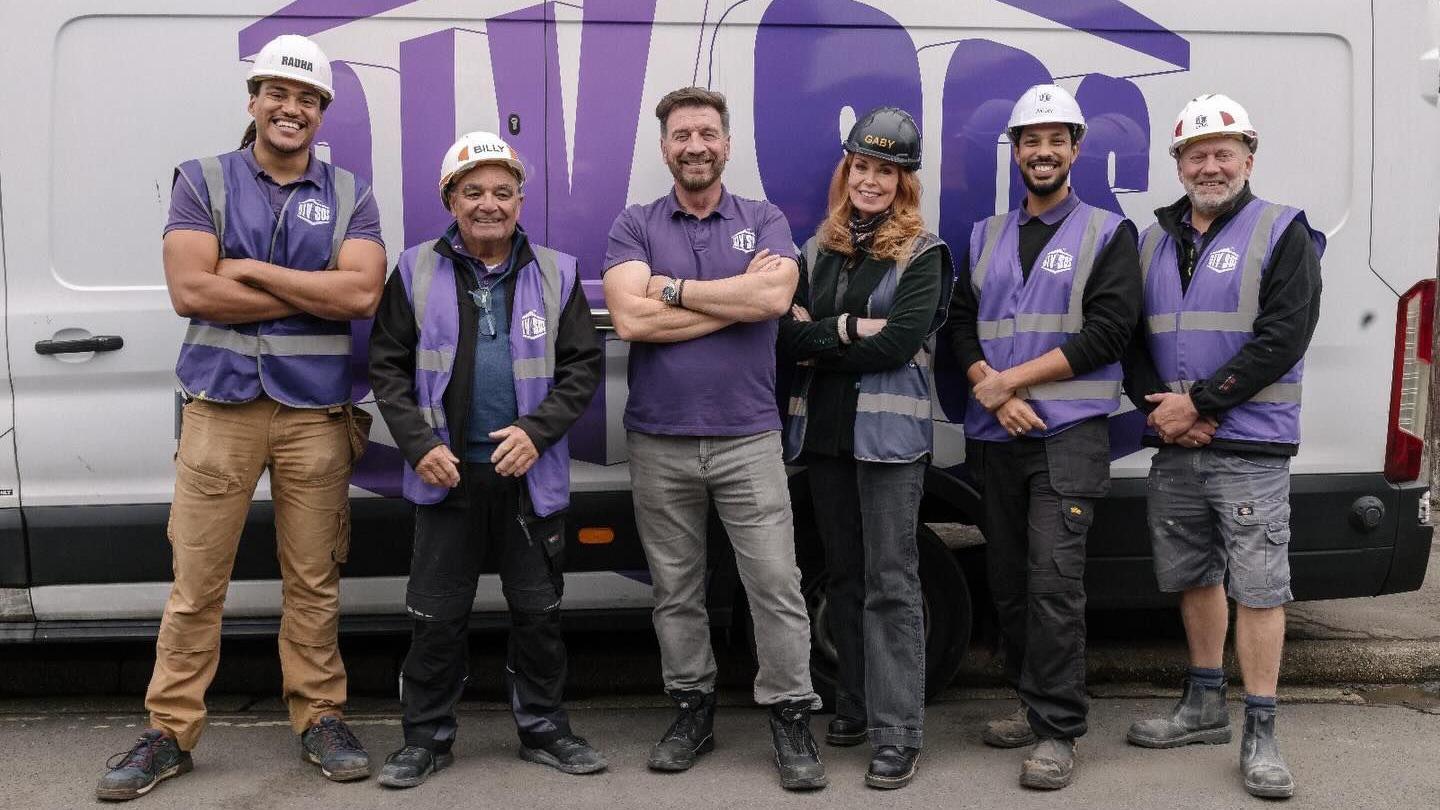 Llun o dîm DIY SOS yn sefyll yn edrych at y camera ac yn gwenu gyda fan gwyn tu ôl iddyn nhw. 6 person sydd, 5 dyn a 3 menyw. I'r chwith ma dyn tal gyda het galed wyn ar ei ben. Mae'n gwisgo crys-t porffor a throwsus melyn. Nesa iddo fe mae dyn arall sy'n gwisgo het galed wyn a siaced du. Nesa mae dyn gyda gwallt brown, crys-t porffor a throwsus llwyd. Yna mae menyw gyda het galed du, siaced du a throwsus du. Nesa iddi hi mae dau ddyn arall. Mae gan y ddwy het galed wyn a siwmper du.