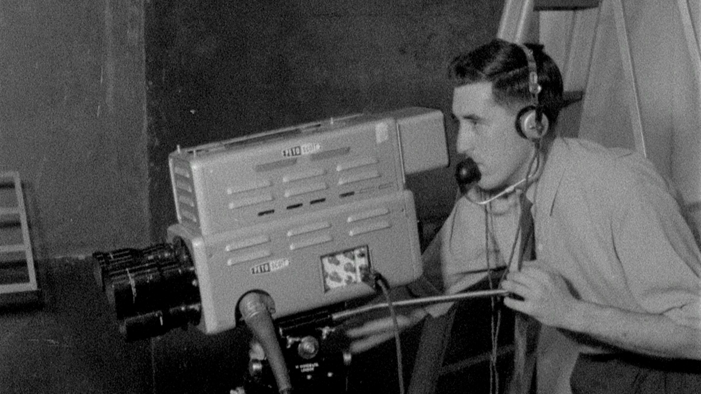 A man operating a television camera in the 1960s. He is wearing a shirt with sleeves rolled up and a tie. He has headphones and a microphone.