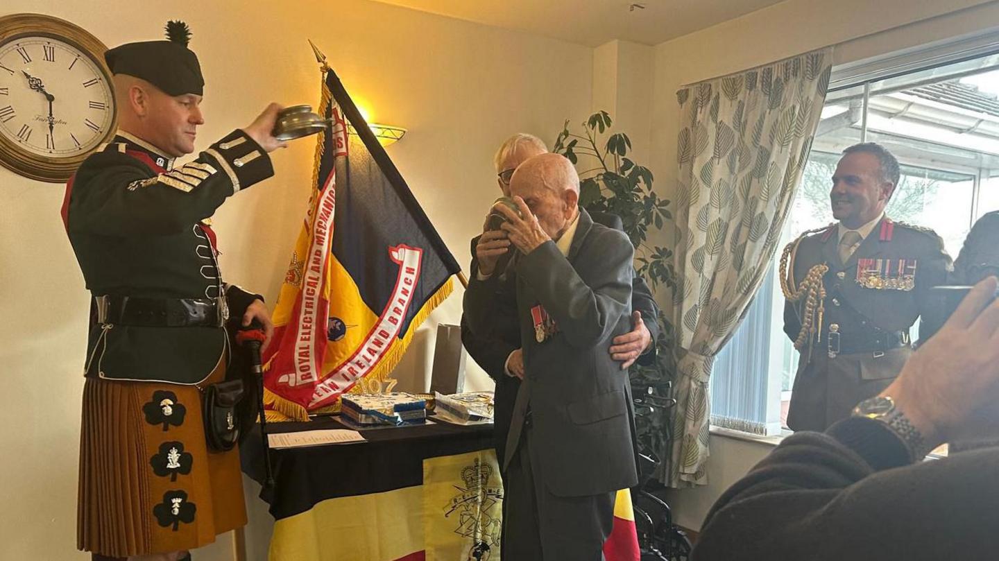 Mr Irwin wearing a suit with his military medals stands up in front of a table which has cake and whiskey sitting on it. He is surrounded by military representatives, and another man has his arms around him. The flag on the table and hanging up is yellow and black, and the military representatives are wearing official uniform.