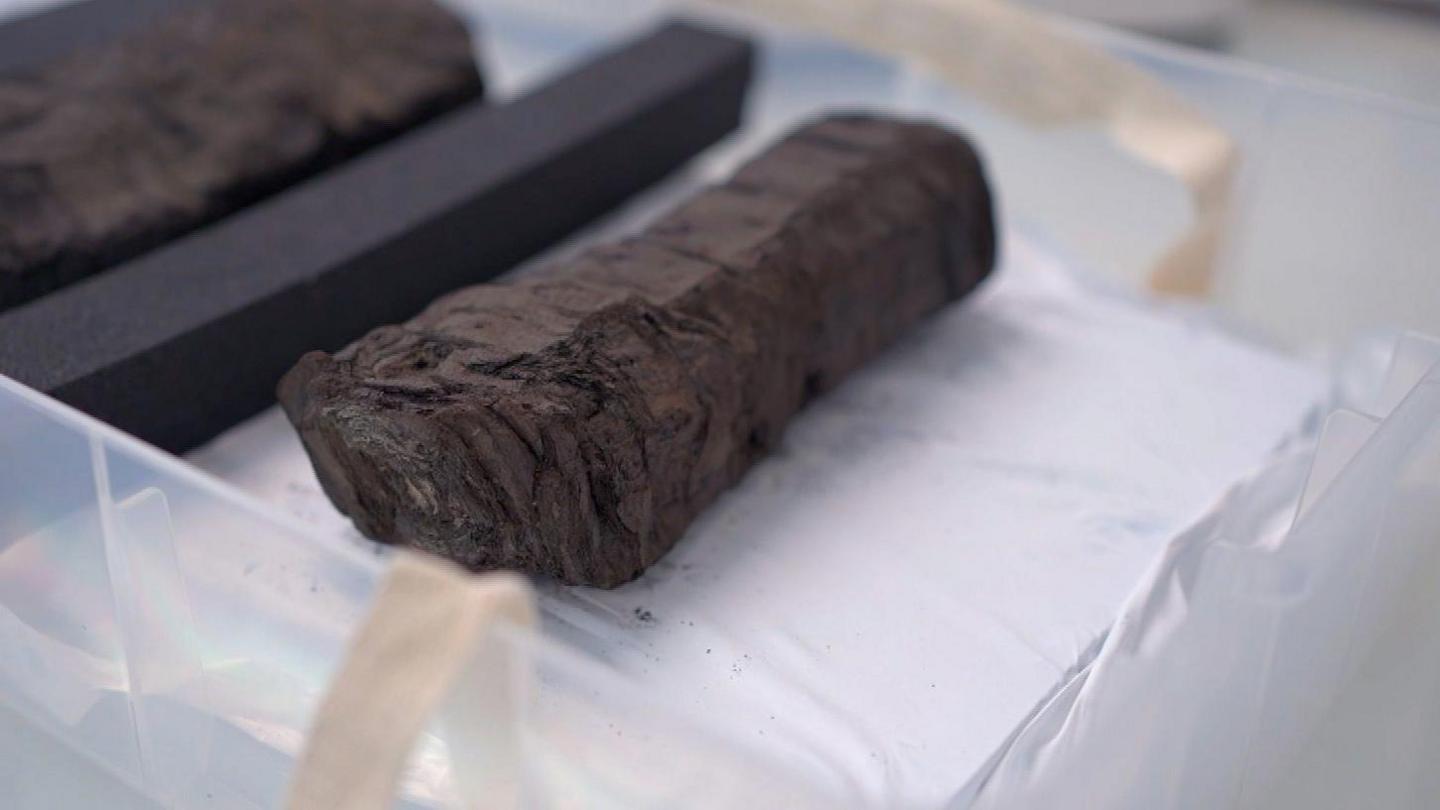 A scroll from ancient city of Herculaneum sitting in a plastic tray on a desk at the Bodleian library. It is about about 15cm long and about 3cm wide. It is dark brown and appears very charred, resembling a lump of charcoal.