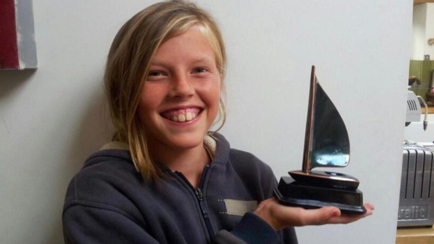 A young girl wearing a navy blue hoodie gives a toothy grin to the camera. Her hair is fair and swept to one side in a low ponytail. In her right hand, which is held across her body, she is holding up a statuete of a sailing yacht. She is posing in front of a plain white wall. In the background is the corner of a toaster. 
