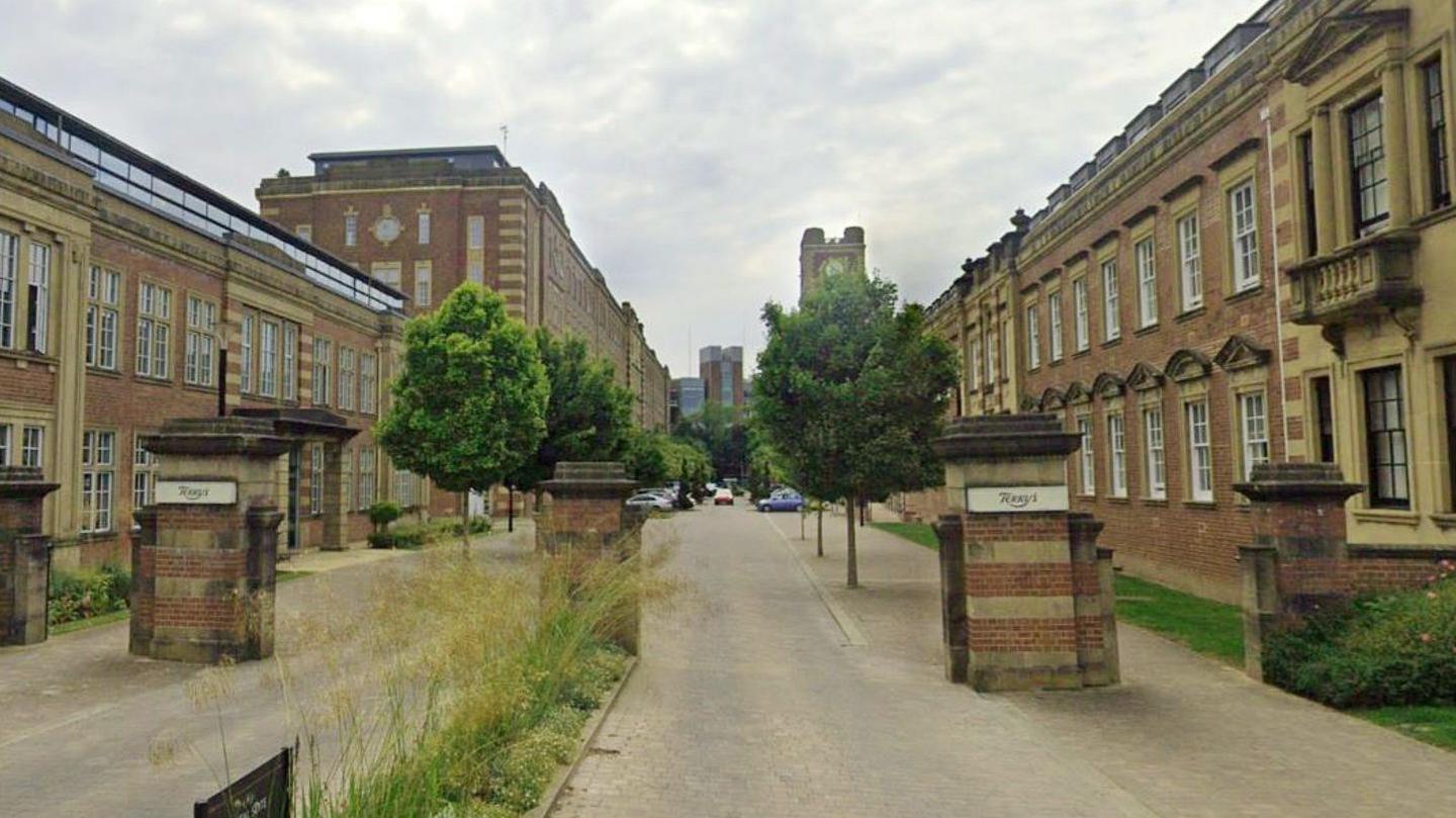 Brick pillars with a plaque with Terry's written at the top of each column mark the entrance to a straight road between two former factory buildings leading to a tall clock tower.