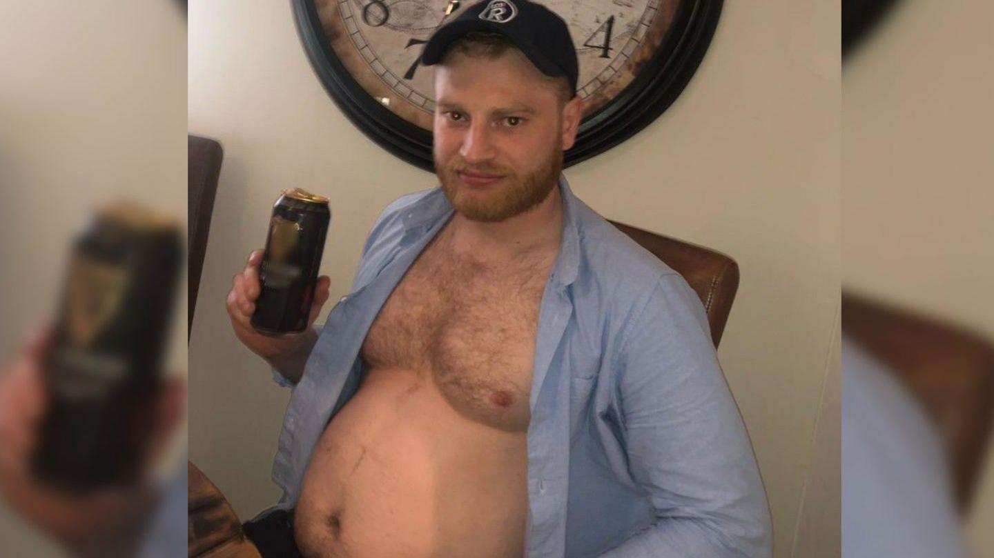 A man sat with a can of beer, his shirt is open to show his stomach.