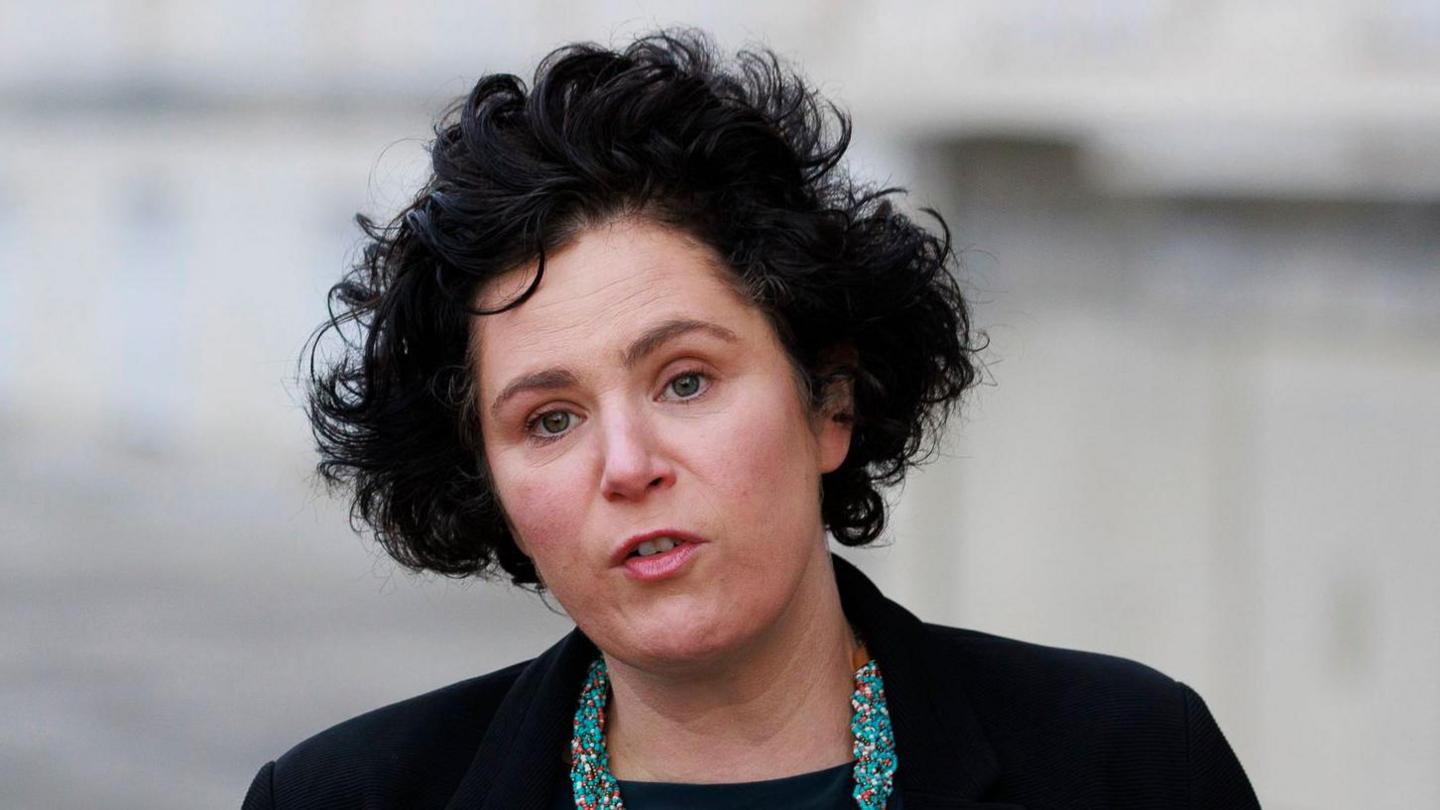 Ms Hanna with short black curly hair speaks to the camera. She is wearing make-up, a black blazer and black top with a blue beaded necklace. The background is blurred.