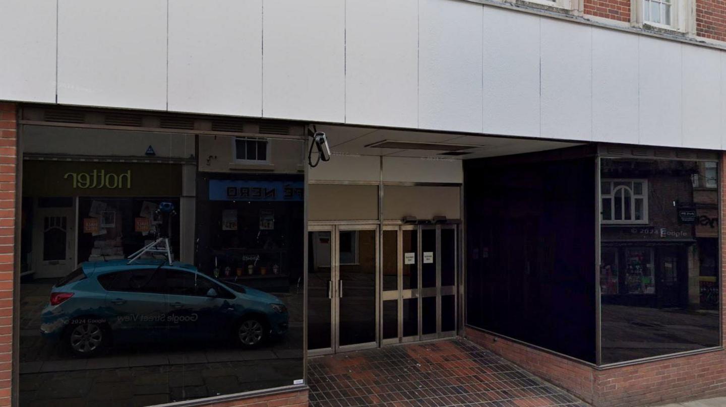 An empty shop. The windows are blacked out and there are two sets of doors in the middle, one of which is automatic. A lone CCTV camera points down towards the entrance.