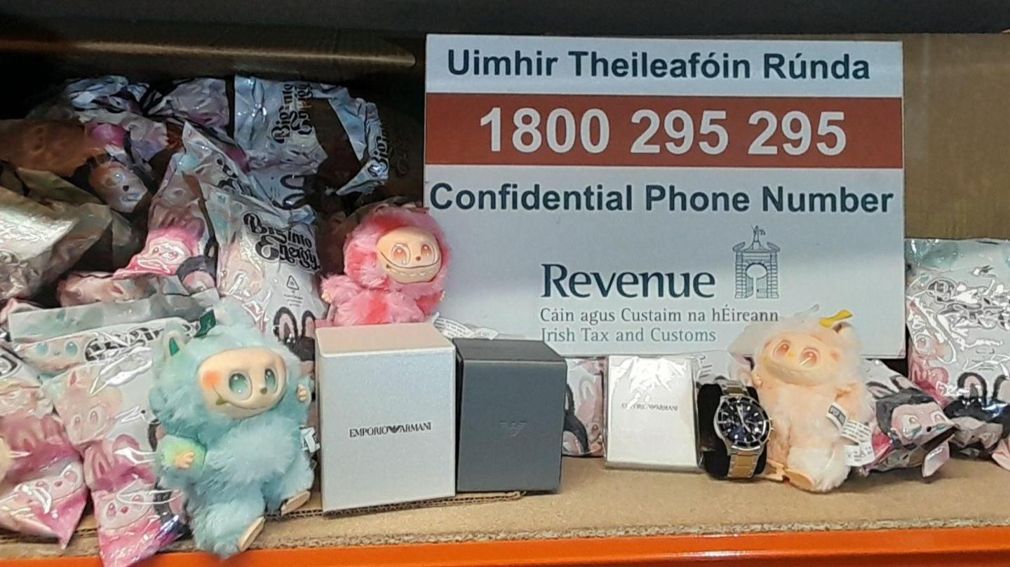 Blue, yellow and pink Labubu dolls next to a sign for Irish Revenue. Watches and their boxes are also present.