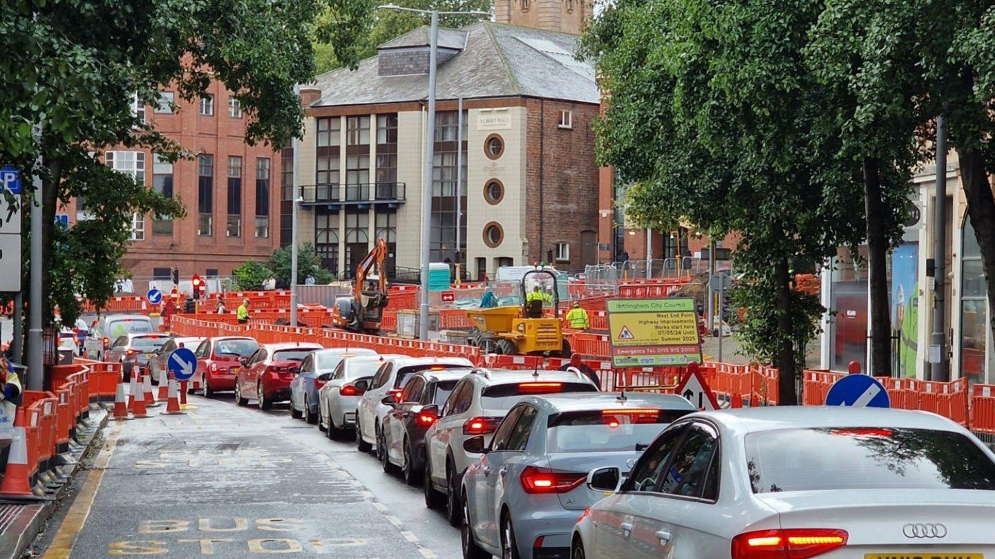 A queue of at least 10 cars displaying red brake lights wait on Upper Parliament Street in Nottingham on the approach to the new T-junction on Maid Marian Way. Construction barriers, traffic cones and building machinery can be seen in the background.