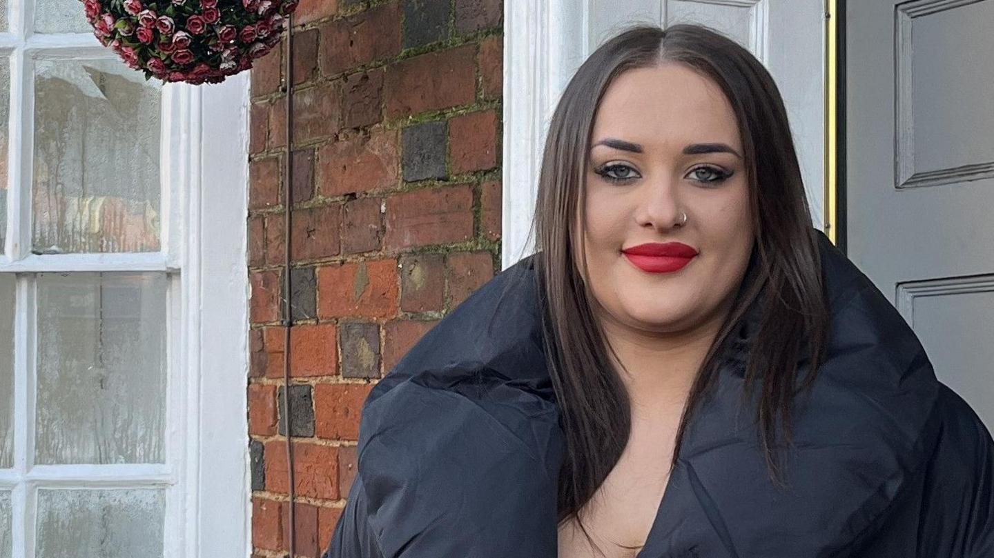 Ellie stands outside her front door which is grey amid traditional brickwork. The row of houses continues behind her, with white bay windows and a sign for the New Inn pub in the distance. Ellie has bright red lipstick and wears a big navy puffer coat. She has straight dark brown hair.