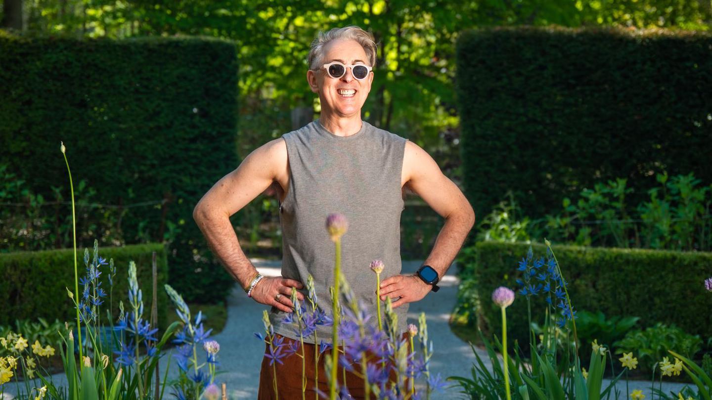 Alan Cumming standing in a garden with a pond behind him. He is wearing white sunglesses and a grey cut off T-shirt. he is standing smiling, with his hands on his hips