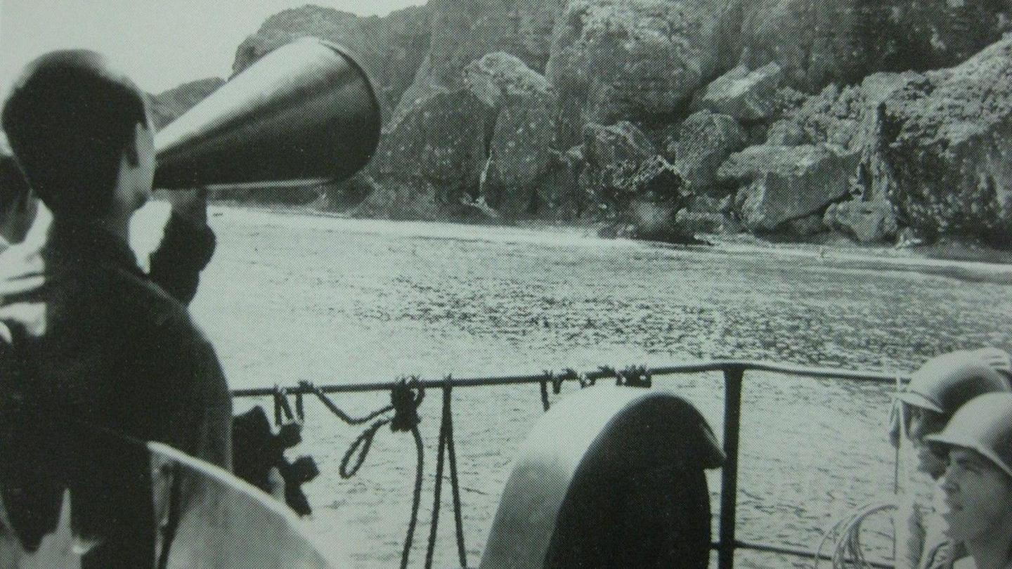 A black and white image of US soldiers in a boat, with one in the front using a loudspeaker. In the distance is an island with a rocky cliff