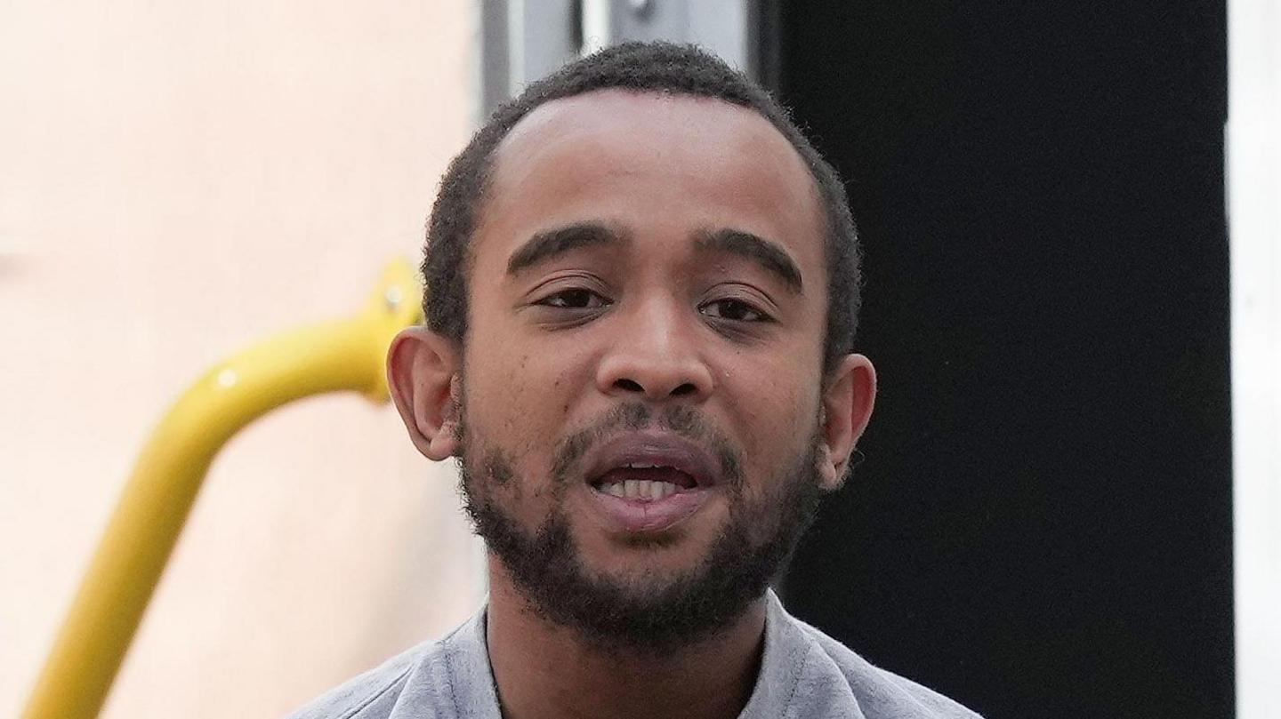 A man with short hair in a grey jumper being lead from a prison van