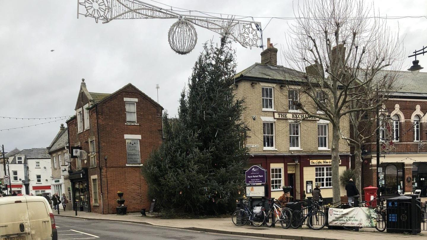 A 9m (30ft) Christmas tree which is leaning to the right in the Market Square, March. Shops are behind it and large decorations hang across the road in front of it in 2023