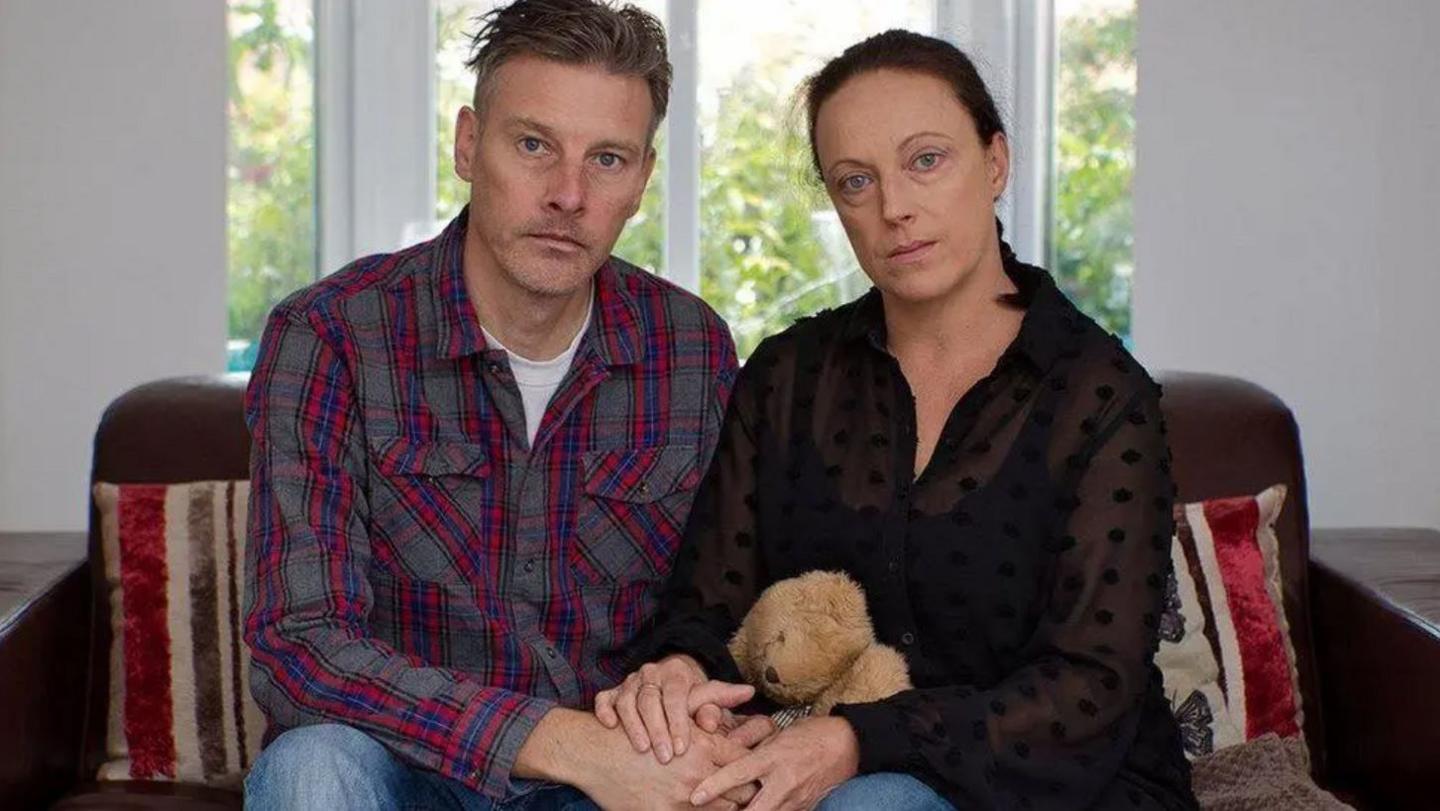 A man with short hair and a blue and red chequed shirt sat beside a woman with long dark hair and a black top and they are both holding a teddy