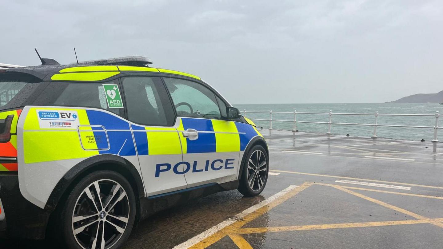 A police car is parked in a car park. In the background the sea can be seen. 