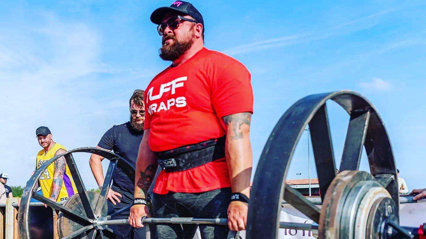 Andrew Reid is wearing a tight red t-shirt, a black cap and sunglasses as he lifts a large bar-bell with metal weights on either end.