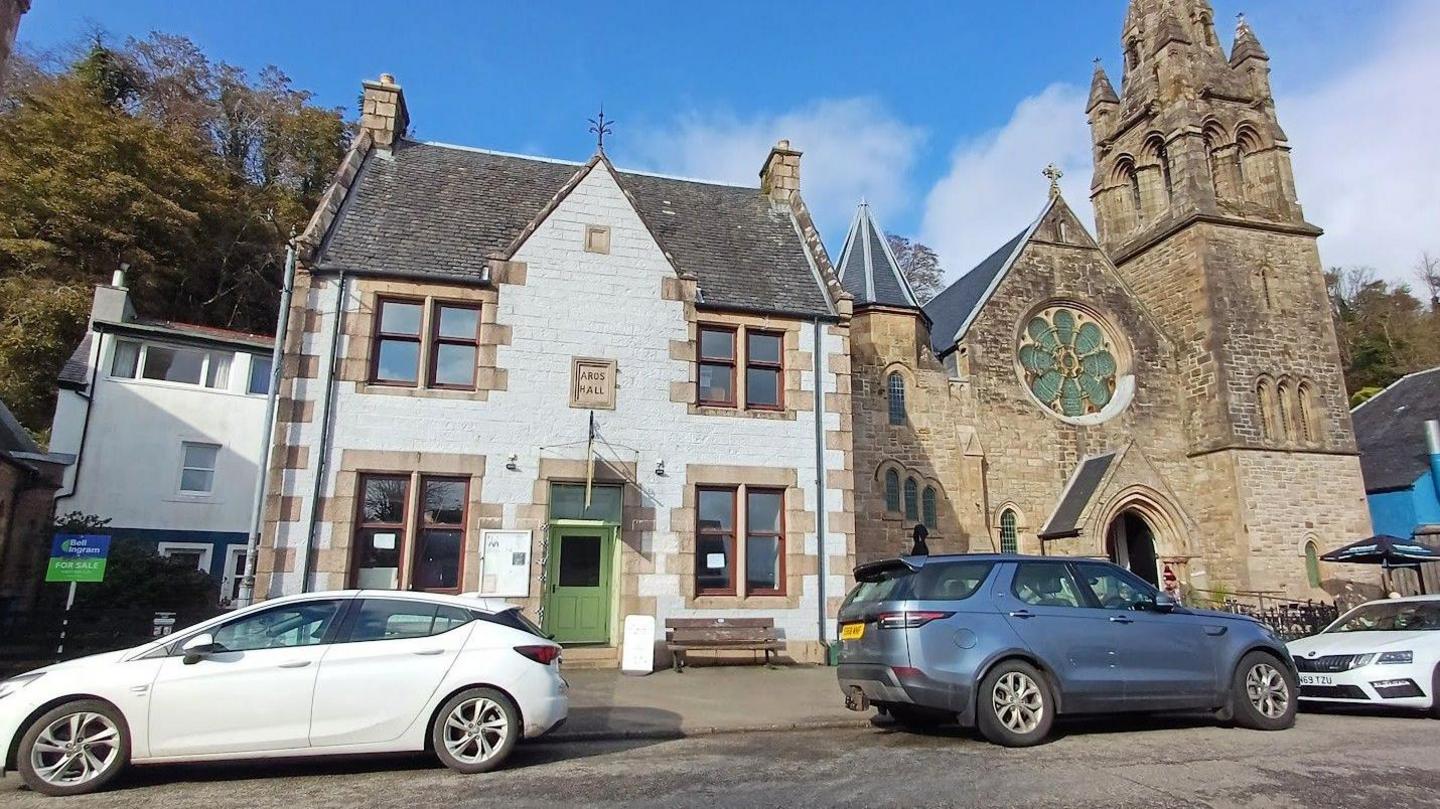 The hall is a small, two-storey building with a green door and square windows. It is next to an old church. Three cars, one blue and two white, are parked on the street outside the buildings.