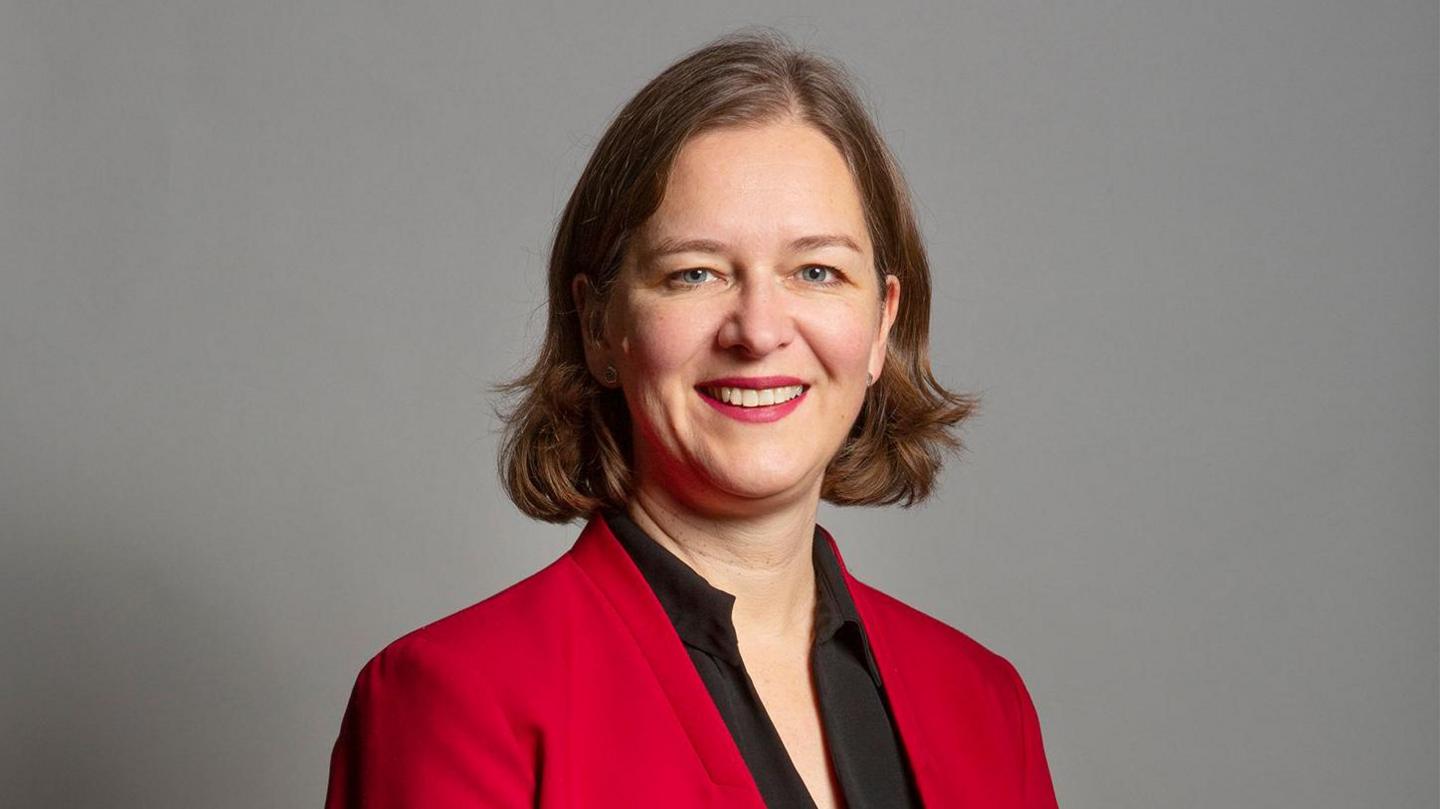 Fleur Anderson wearing a red suit with a black top in front of a grey background