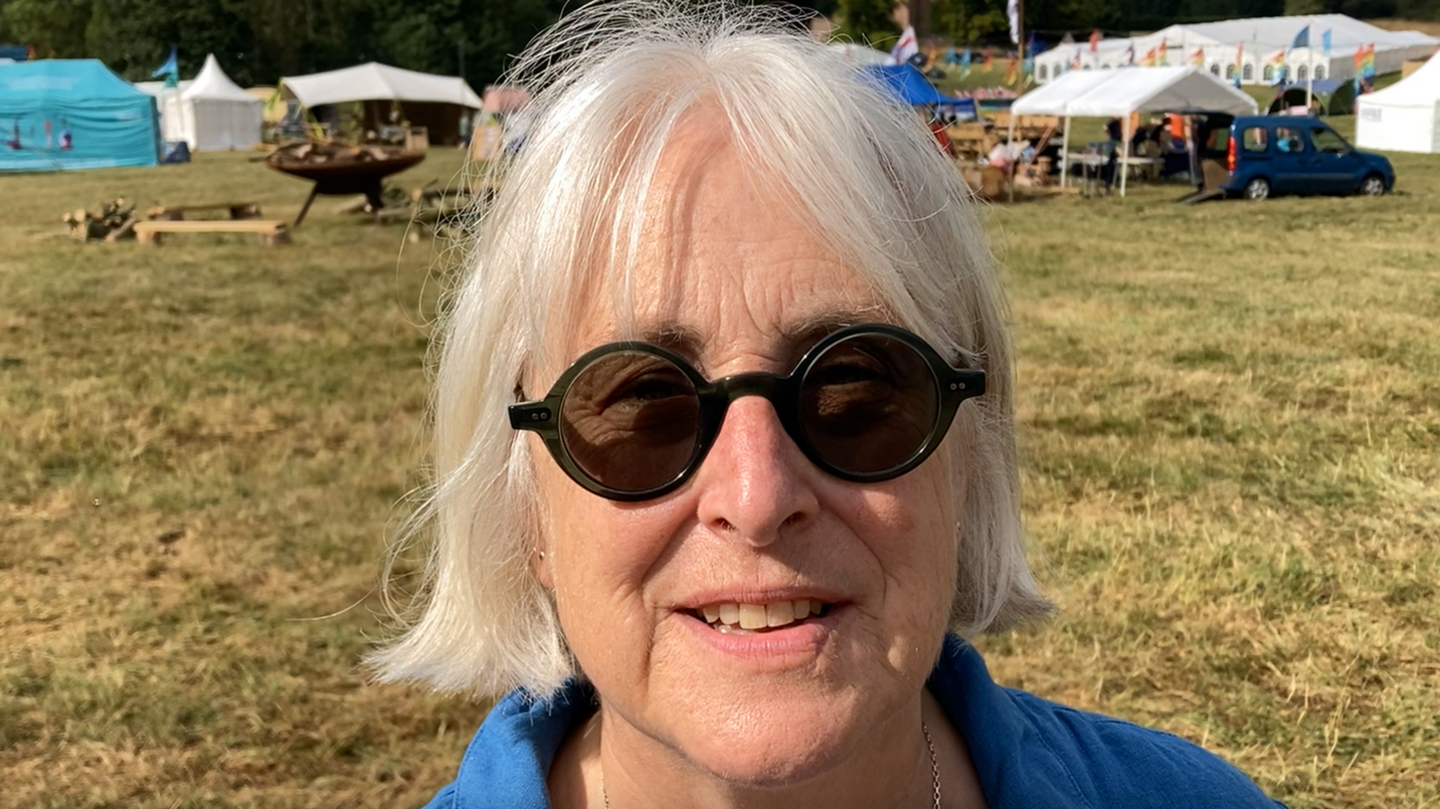 Becky Marshall-Potter photographed outside, she is wearing a blue jacket and dark sunglasses. Marquees, tents and cars are behind her. 