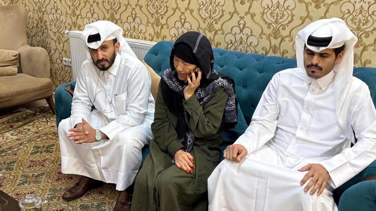 Faye Hall sits on a blue sofa in between two men wearing white in a plush room with patterned yellow wallpaper and carpeting. She wears a long dark green dress with long sleeves and a black and white head scarf and is speaking into a mobile phone.