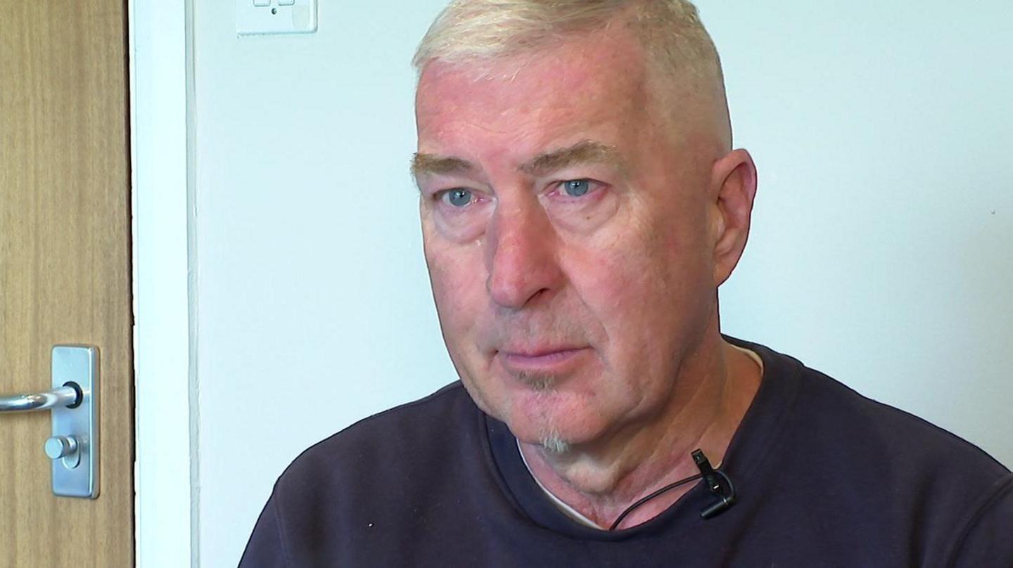 Rob Hobson looking away from the camera. He is wearing a black jumper with a white t-shirt underneath. He is sitting indoors, in front of a white painted wall and a wooden door.