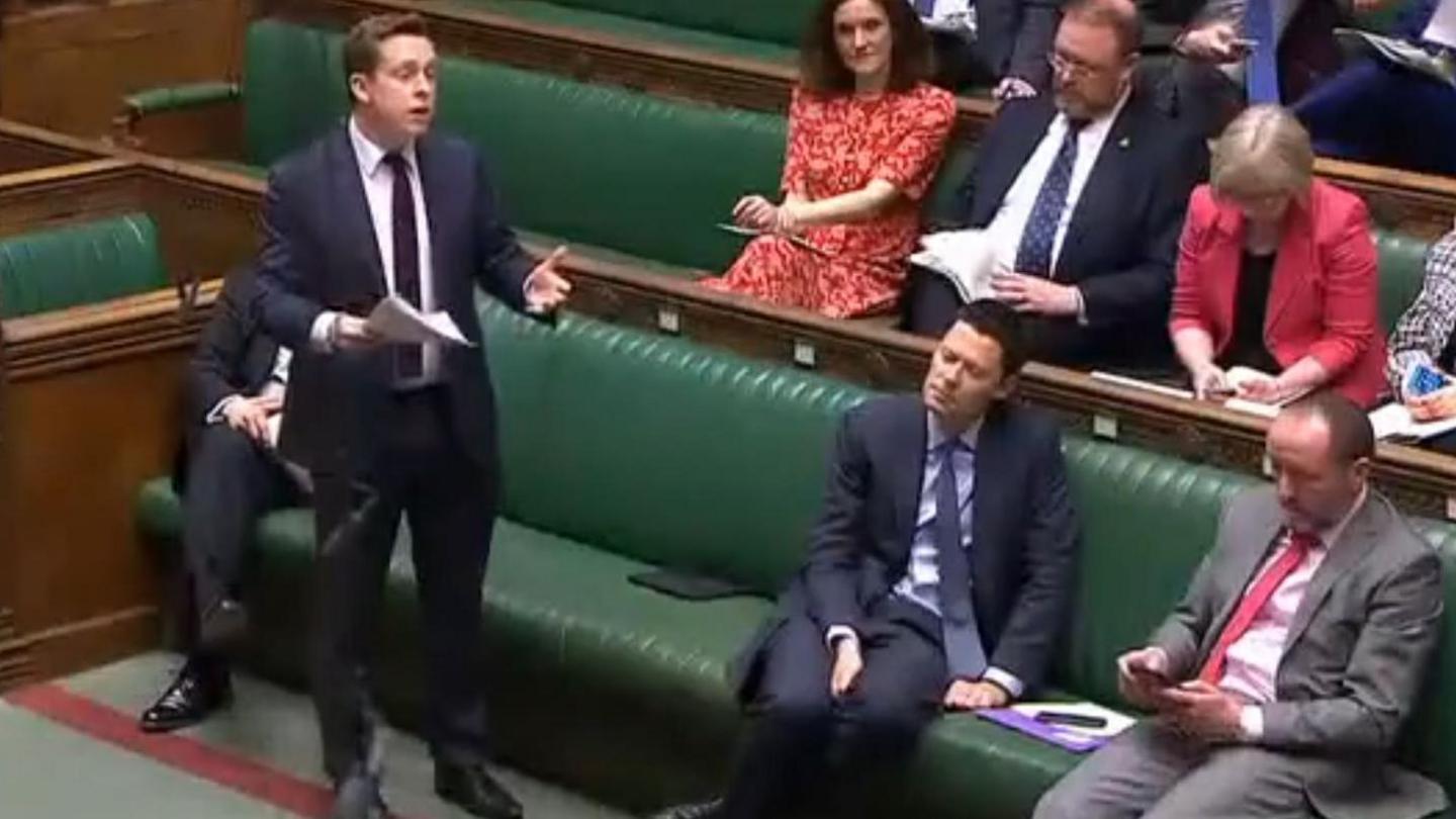 Tom Pursglove with short fair hair standing in front of the green benches in the House of Commons. He is holding a sheaf of papers.  An MP sitting close by appears to be looking at his phone.