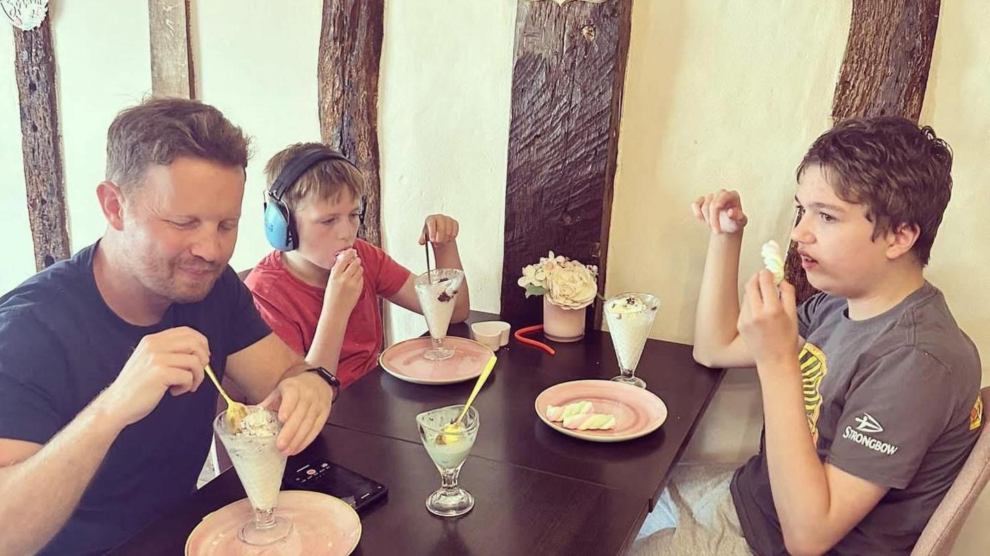 James, Jude and Tommy are at a cafe eating ice creams. James and Tommy are sat on side of the table with Tommy wearing headphones. Jude is sitting opposite Tommy. There are four ice creams, all in sundae type glasses on the table, but one looks like it has already been finished.