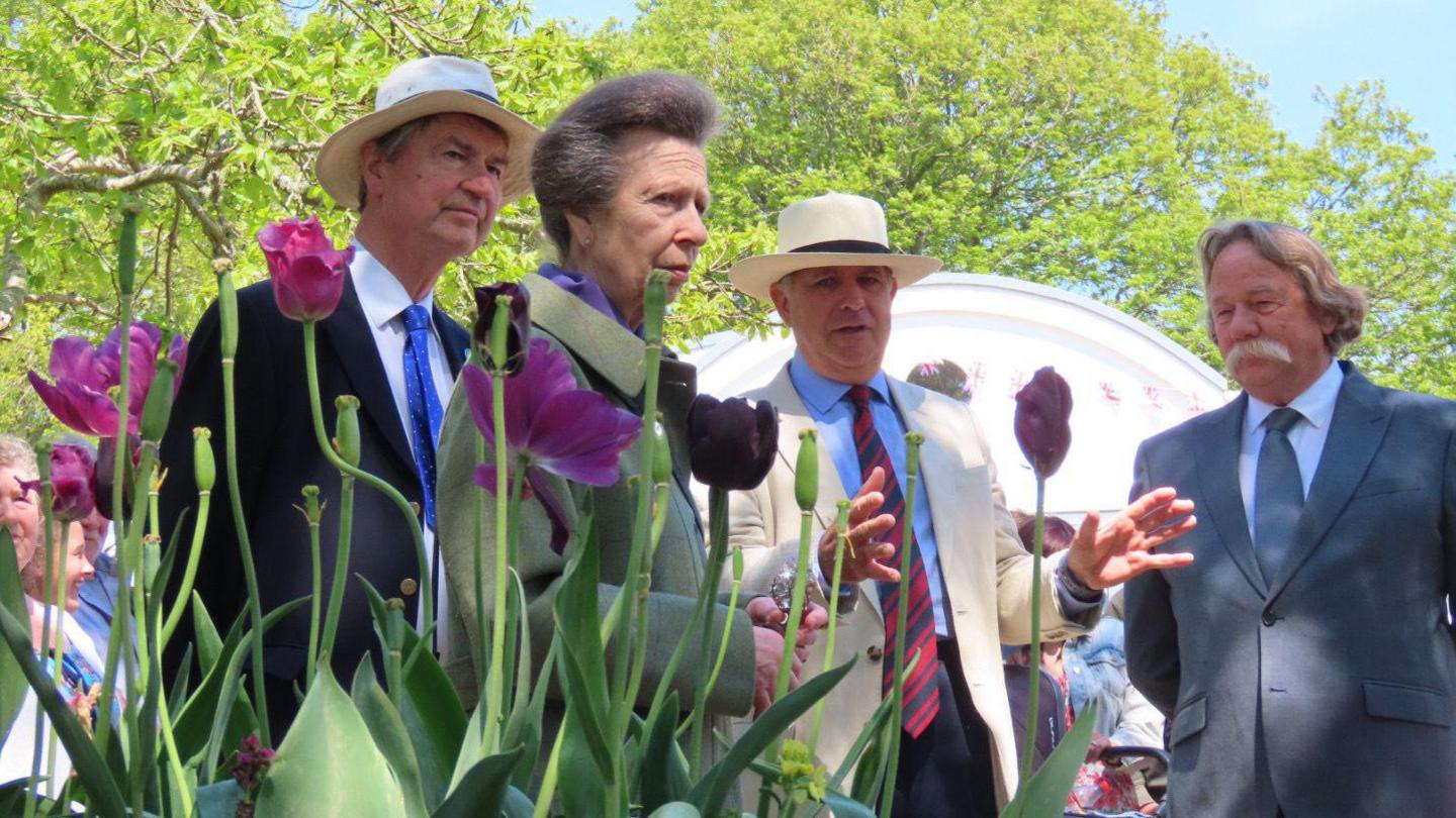 Princess Anne standing outside listening as a man talks to her. There are two other man standing with her and behind them are spectators and a large tent. In the foreground there are tulips.