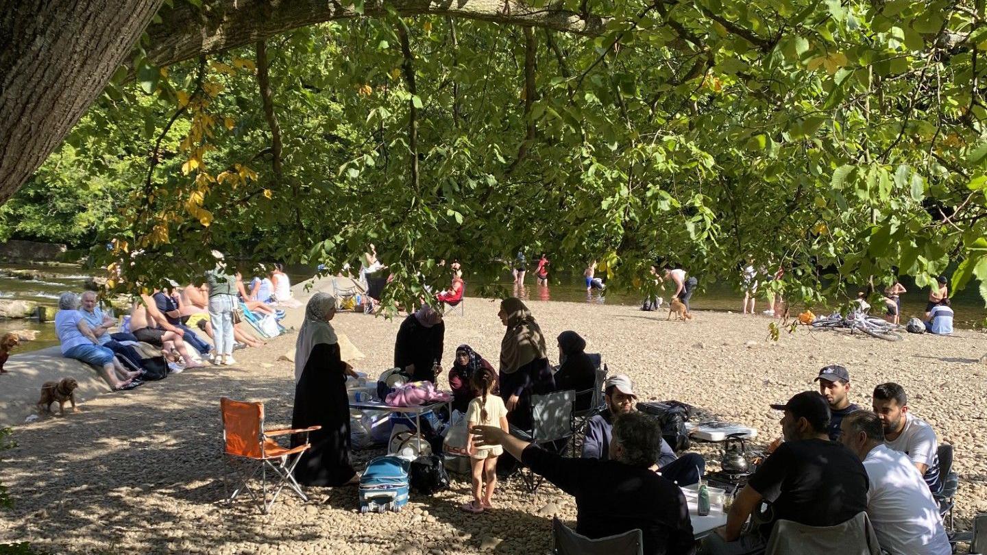 Groups of people sit on a river bank and paddle in the water. It is a sunny day and the site is shielded by trees. 