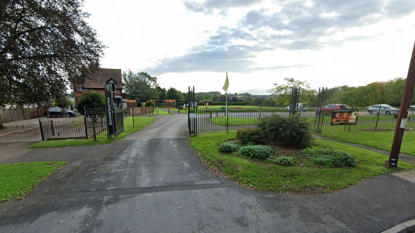 The entrance of a park with black gates and fencing. A green flag is flying inside the park. Trees can be seen in the distance.