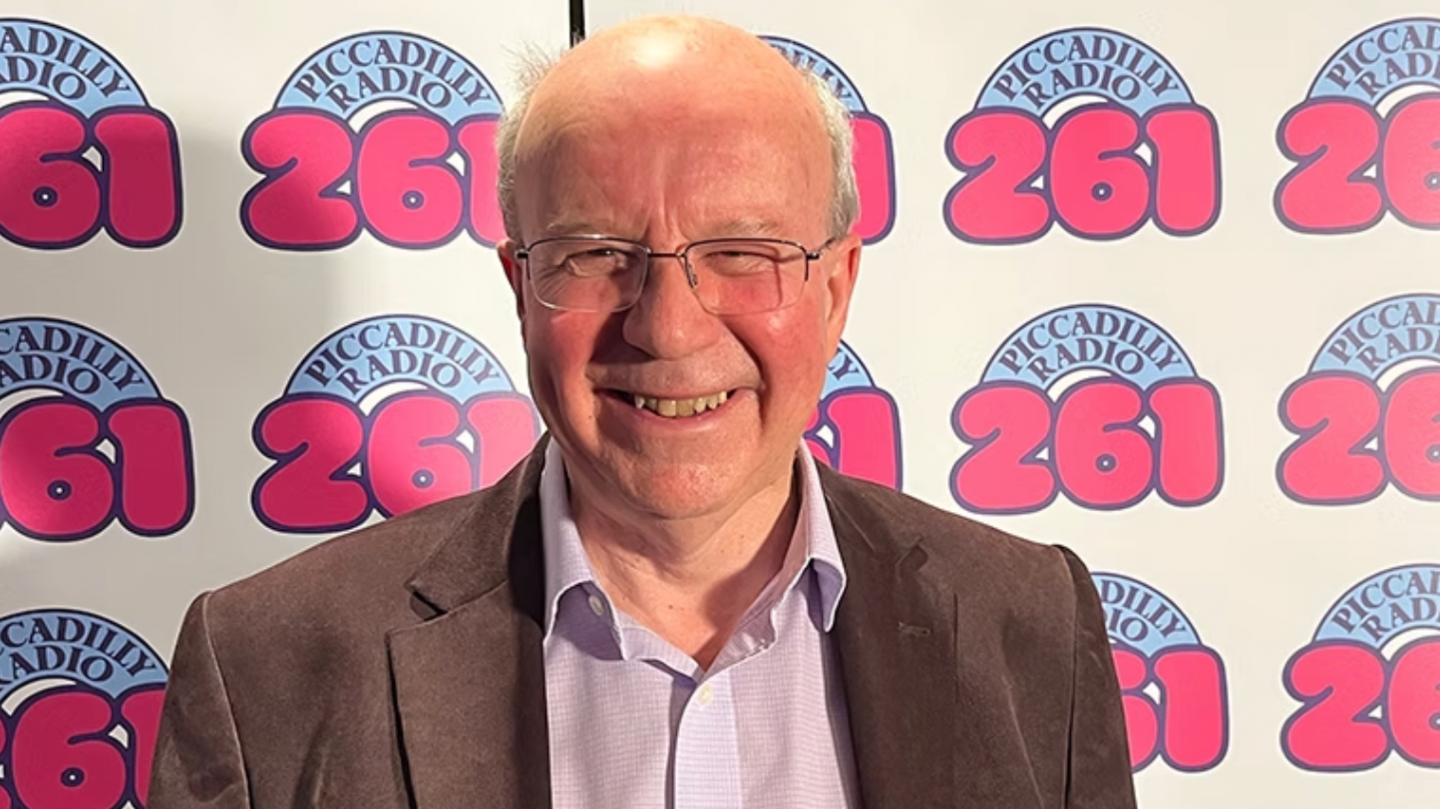Jim Hancock stands in front of the camera and smiles. He's wearing glasses and a brown suit behind a backdrop of Piccadilly Radio's logo.