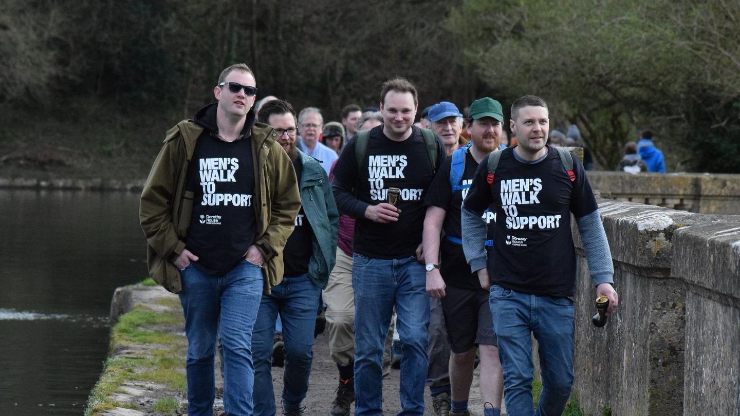 A group of men walking by a river. Four of the men are wearing black T-shirts which say Men's Walk to Support