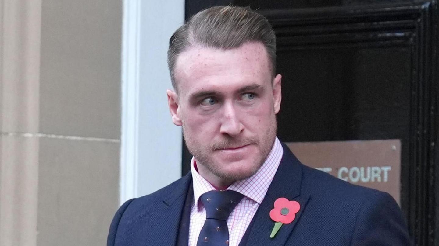 Stuart Hogg grimacing as he comes out of court. He is wearing a suit and tie with a poppy attached to his jacket.