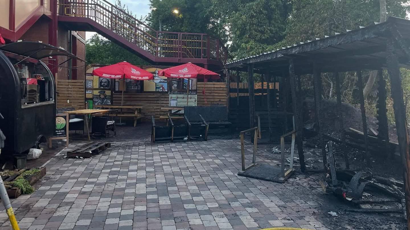An outdoor seating area for a bar at a railway station. The wooden structure on the right where people would have sat is burnt down. Tables and chairs are spread across the floor and are broken, covered in black ash. A staircase and bridge connecting two railway platforms can be seen above.
