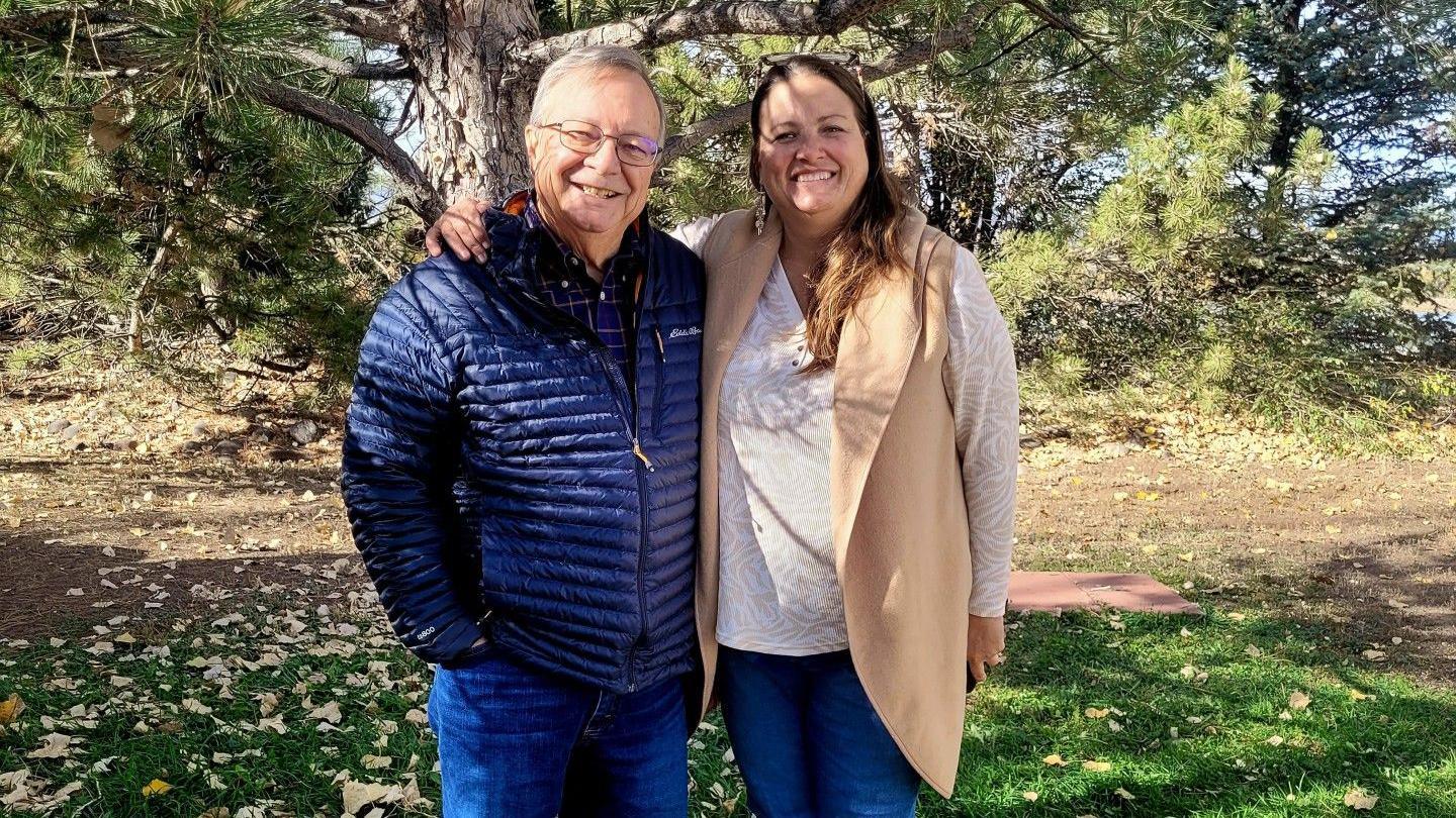 A dad and a daughter are seen hugging and posing for a picture in a park in front of a tree with fallen leaves.