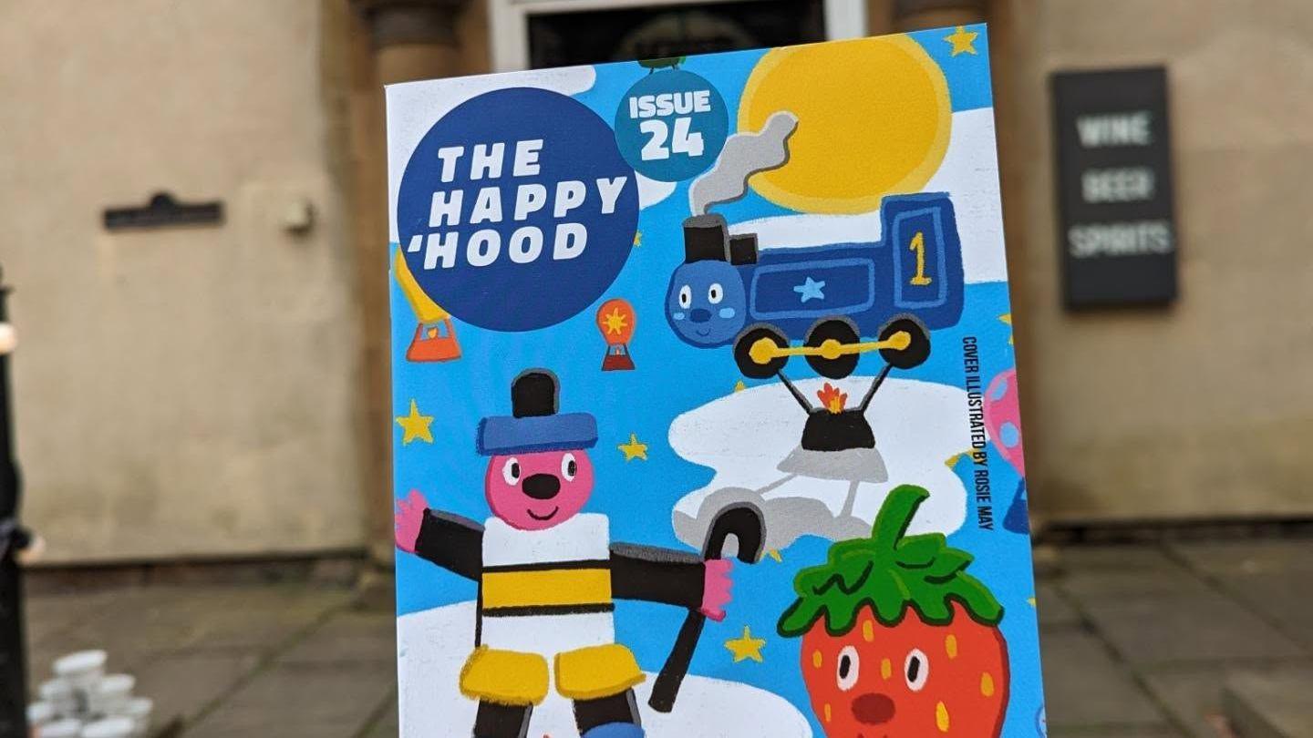 A blue magazine called The Happy Hood being held up. The front cover features various drawings including a train, a man made of sweets and a stawberry.