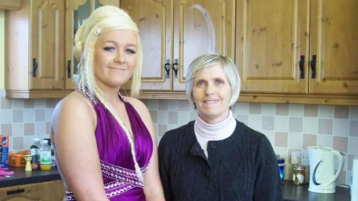 A woman with blonde hair, wearing a purple dress, standing beside a woman with short grey hair, wearing a black jumper. They are standing in a kitchen. 