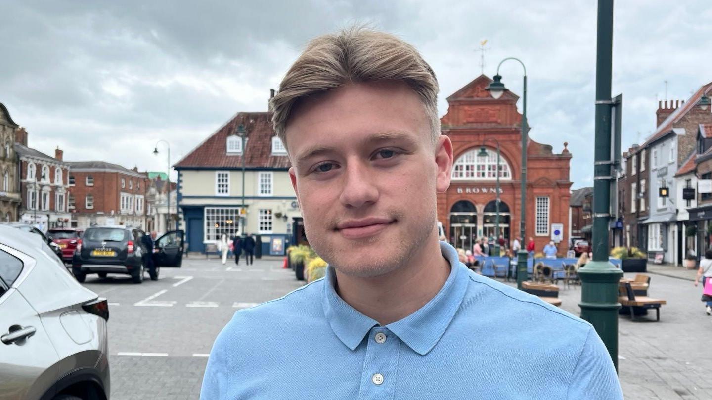 Dominic Brown, a man in his 20s standing in the busy market square wearing a blue top, with cars and people in the background 