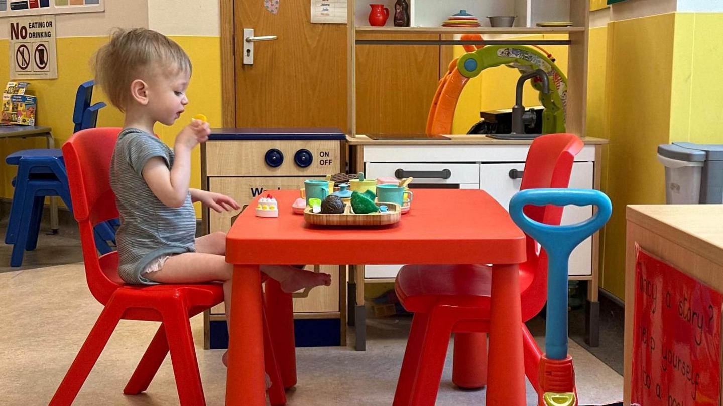 A two-year-old boy sat on a small red chair pulled up next to a small red table. He is playing with toy food and in the background is a toy kitchen.