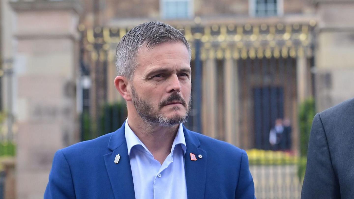 A man in a blue suit, wearing a blue shirt, and with a black and grey beard and short greying hair. He is standing in front of a building.