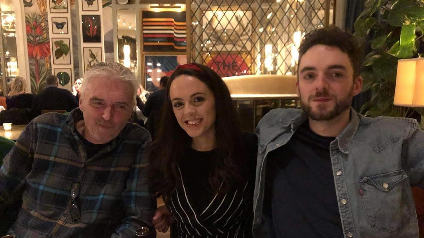 Three people sat next to each other in a restaurant. Left, a man with short white hair wearing a checked shirt. Middle, a woman with brown hair wearing a black dress with white stripes. Right, a man with short brown hair and a short beard wearing a blue t-shirt and grey denim overshirt. They are sat in a colourful room with lots of photos on the walls.