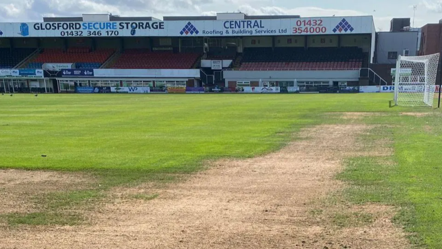 A football pitch with dry brown patches. There is a white goal net and football seat stands.