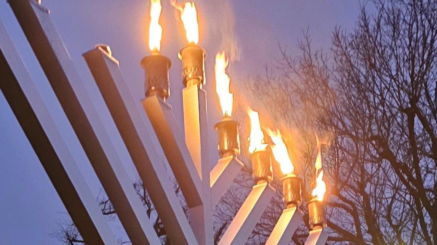 A metal candelabrum with eight branches and one central one set against a darkening sky with a tree in the background. Five of the candles on the branches are lit along with the central candle.