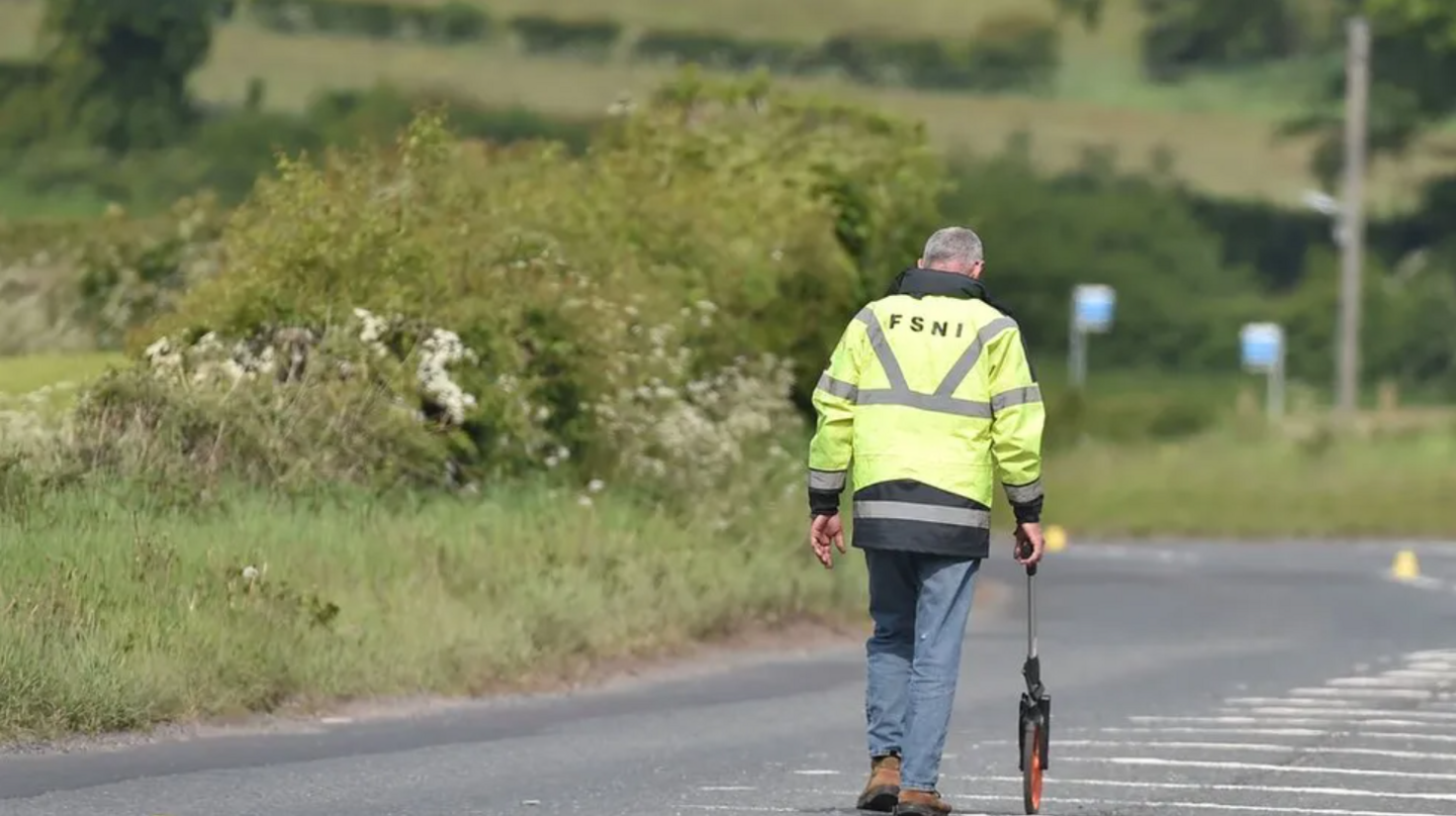 A man wearing a hi-vis jacket with the letters FSNI on the back walking down a road, carring a measuring device