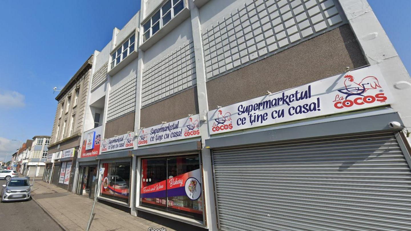 The building of 172-182 High Street, which has white panelling on the upper floors, with small square tiling. On the ground floor is Romanian supermarket La Cocos, which has blue writing, the outline of a chicken at either end and a white background